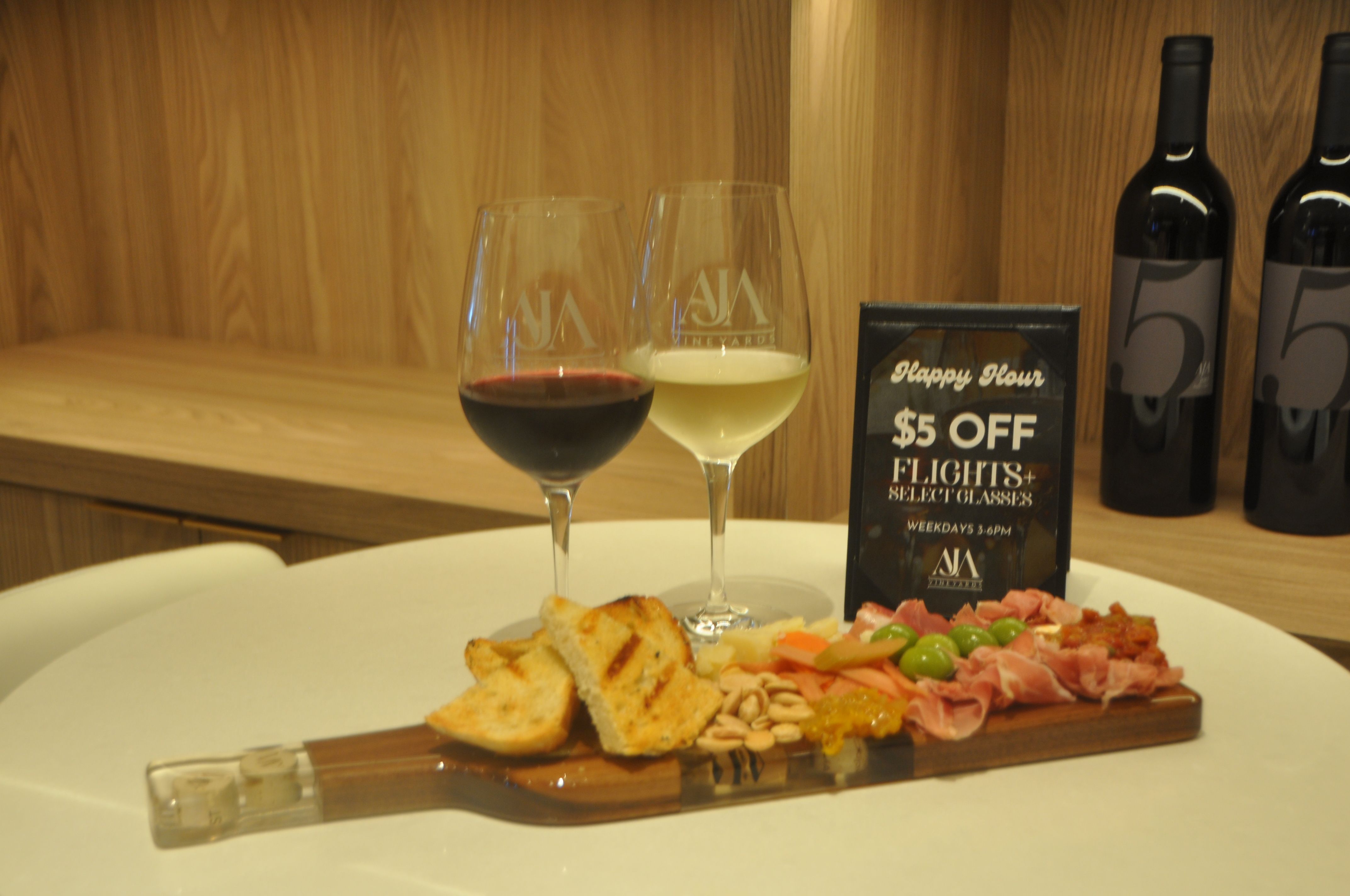 Two wine glasses (red and white) with wine bottles in the background, a charcuterie board of grilled bread, cured meats, olives, nuts and preserves, and a small sign advertising $5 off flights for happy hour on a wooden table.