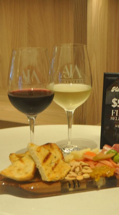 Two wine glasses (red and white) with wine bottles in the background, a charcuterie board of grilled bread, cured meats, olives, nuts and preserves, and a small sign advertising $5 off flights for happy hour on a wooden table.