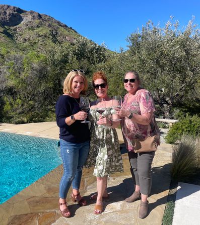 Three friends toasting with red wine by a bright blue pool on a stone patio, framed by olive trees and a sunlit hillside under a clear blue sky.