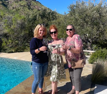 Three friends toasting with red wine by a bright blue pool on a stone patio, framed by olive trees and a sunlit hillside under a clear blue sky.