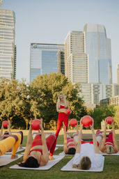 Sunlit outdoor fitness class in an urban park with downtown skyline: instructor in red holding a red exercise ball while participants lie on mats lifting red balls.
