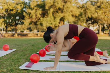 Person in burgundy activewear doing a knee-to-elbow core move on a yoga mat during an outdoor fitness class in a sunny park with red exercise balls nearby