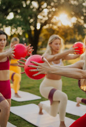 Group of women in an outdoor fitness class doing lunges on yoga mats in a sunlit park, holding bright red exercise balls and wearing resistance bands.