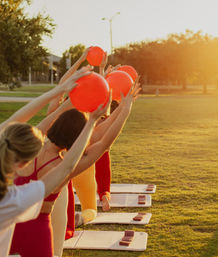 Group doing outdoor yoga in a sunlit park at sunset, kneeling on mats and lifting bright red exercise balls overhead during a golden-hour fitness class.