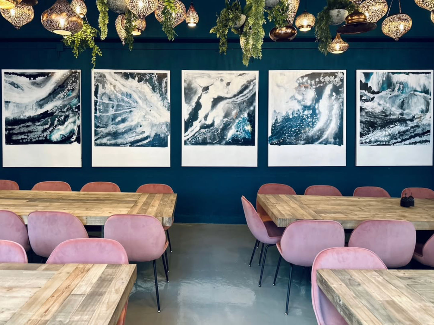 Modern cafe interior with a teal accent wall showcasing five large abstract ocean paintings, rustic wooden communal tables, pink velvet dining chairs, and decorative hanging brass lanterns with trailing greenery.