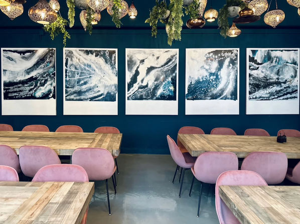 Modern cafe interior with a teal accent wall showcasing five large abstract ocean paintings, rustic wooden communal tables, pink velvet dining chairs, and decorative hanging brass lanterns with trailing greenery.