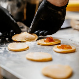 Gloved hands in an artisan bakery sealing jam-filled round pastry cookies on a floured stainless steel prep surface