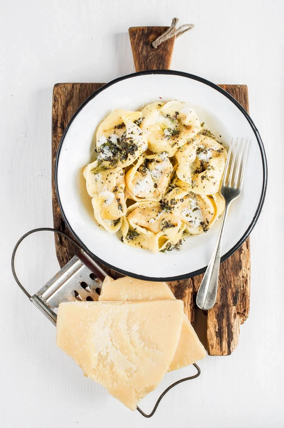 Top-down view of Italian cheese tortellini in creamy herb butter sauce in a white enamel bowl on a rustic wooden board, fork and Parmesan wedges with grater beside it