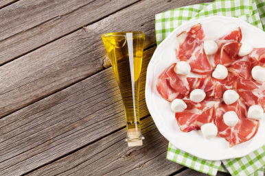 Italian antipasto: white plate of thin prosciutto slices topped with small bocconcini mozzarella balls beside a slender glass olive oil bottle on a green checked napkin and rustic wooden table.