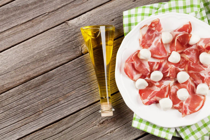 Italian antipasto: white plate of thin prosciutto slices topped with small bocconcini mozzarella balls beside a slender glass olive oil bottle on a green checked napkin and rustic wooden table.