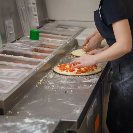 Hands of kitchen staff topping pizza crusts with diced tomatoes and shredded cheese at a stainless-steel pizza prep station with ingredient bins, flour-dusted counter