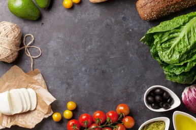 Bright Mediterranean ingredients on a dark slate countertop: cherry tomatoes on the vine, sliced mozzarella, avocado, romaine lettuce, black olives, pesto, olive oil and seeded bread arranged around an empty center.
