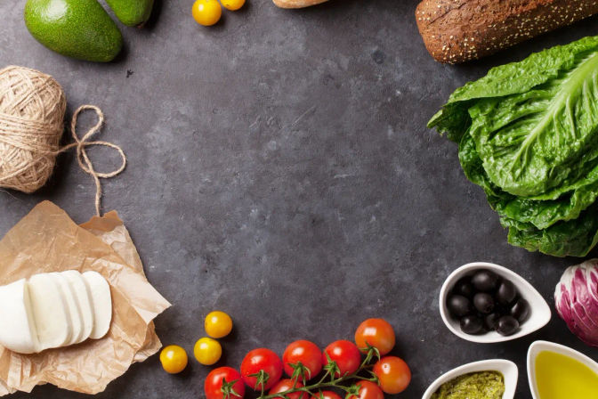 Bright Mediterranean ingredients on a dark slate countertop: cherry tomatoes on the vine, sliced mozzarella, avocado, romaine lettuce, black olives, pesto, olive oil and seeded bread arranged around an empty center.
