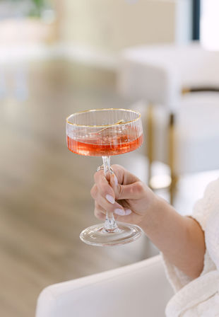 Hand with manicured nails holding a gold-rimmed coupe glass filled with a pink cocktail and stirrer in a bright modern lounge setting