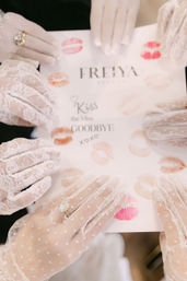 Playful bridal-shower scene with several hands in vintage white lace gloves and rings holding a card splattered with colorful lipstick kiss marks and a handwritten 'Kiss the Miss Goodbye' message.