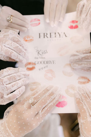 Playful bridal-shower scene with several hands in vintage white lace gloves and rings holding a card splattered with colorful lipstick kiss marks and a handwritten 'Kiss the Miss Goodbye' message.