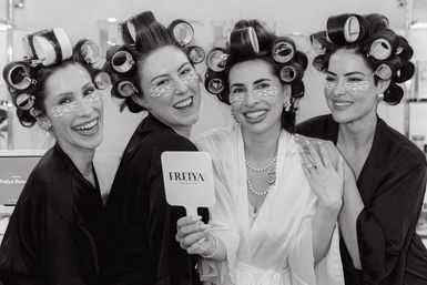 Black-and-white photo of four women in hair rollers and under-eye patches, wearing robes and laughing together while holding a handheld mirror during beauty prep.