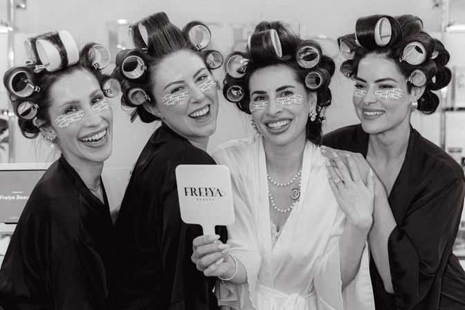 Black-and-white photo of four women in hair rollers and under-eye patches, wearing robes and laughing together while holding a handheld mirror during beauty prep.