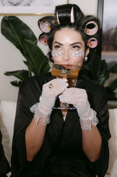 Woman in black robe with pink hair rollers and printed under-eye patches sipping a cocktail from a coupe, wearing white lace gloves and a ring in an at-home getting-ready scene with houseplants