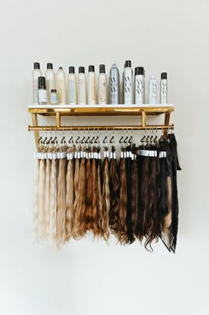 Salon wall display with long wavy hair extensions and color swatches hanging from a brass rack beneath a shelf of haircare bottles, gradient from platinum blonde to deep brown.
