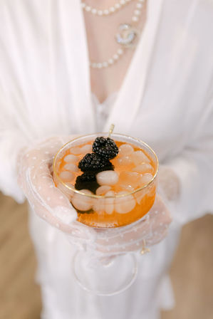Stylish orange cocktail in a gold-rim coupe with lychee pearls and blackberry garnish, held by hands in sheer dotted gloves and a pearl necklace.