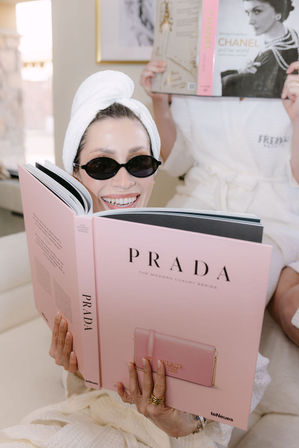 Smiling woman in a towel turban and black sunglasses lounging in a robe, reading a large pink luxury fashion coffee-table book during an at-home spa day while a friend in a white robe reads a fashion book in the background.