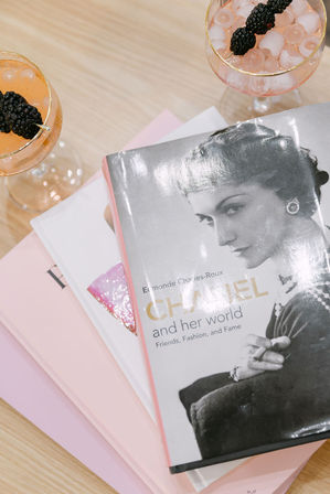 Stack of pastel pink coffee-table books topped by a glossy black-and-white fashion biography, beside two sparkling pink cocktails garnished with blackberries on a light wood table.