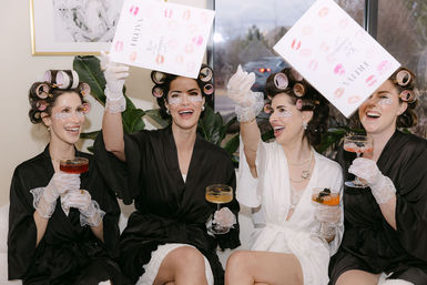 Bridal party getting-ready celebration: four women in satin robes and hair rollers, wearing under-eye patches and lace gloves, laughing and toasting with champagne coupes while tossing wedding-day cards.