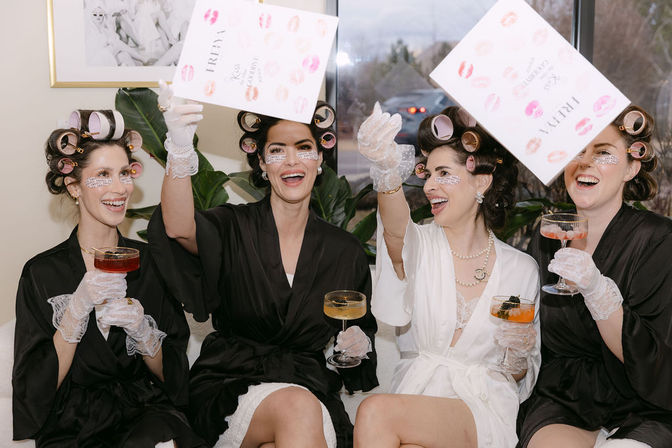 Bridal party getting-ready celebration: four women in satin robes and hair rollers, wearing under-eye patches and lace gloves, laughing and toasting with champagne coupes while tossing wedding-day cards.