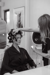 Salon beauty prep: client in a satin robe with hair in rollers having makeup brushed on by a stylist holding a compact; framed cat portrait on the wall.