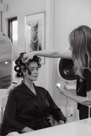Salon beauty prep: client in a satin robe with hair in rollers having makeup brushed on by a stylist holding a compact; framed cat portrait on the wall.