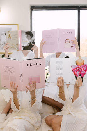 Three people in white bathrobes lounging on a sofa, each holding oversized pink luxury fashion coffee-table books over their faces in a bright sunlit room — stylish spa-day vibe.