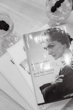 Glossy black-and-white fashion coffee-table book with a vintage portrait, stacked books and two cocktail glasses garnished with blackberries on a light wood table.