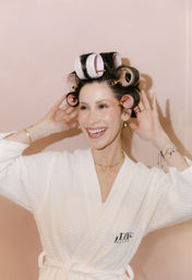 Smiling woman in a white spa robe wearing large pink and white hair rollers while styling dark hair against a soft blush-pink background