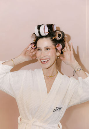 Smiling woman in a white spa robe wearing large pink and white hair rollers while styling dark hair against a soft blush-pink background