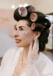 Close-up of woman in salon beauty prep: pink hair rollers, printed under-eye patches, pearl necklace, lace glove and crystal earrings for vintage glam.