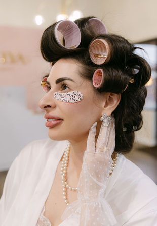 Close-up of woman in salon beauty prep: pink hair rollers, printed under-eye patches, pearl necklace, lace glove and crystal earrings for vintage glam.