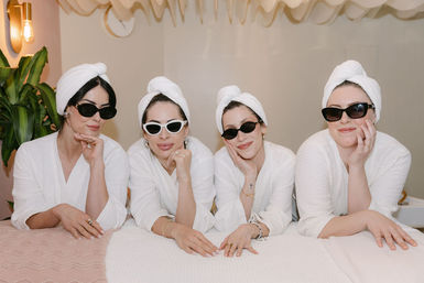 Four women in white robes and towel turbans wearing sunglasses lean on a spa treatment table, smiling in a bright indoor spa room with plants and soft warm lighting.
