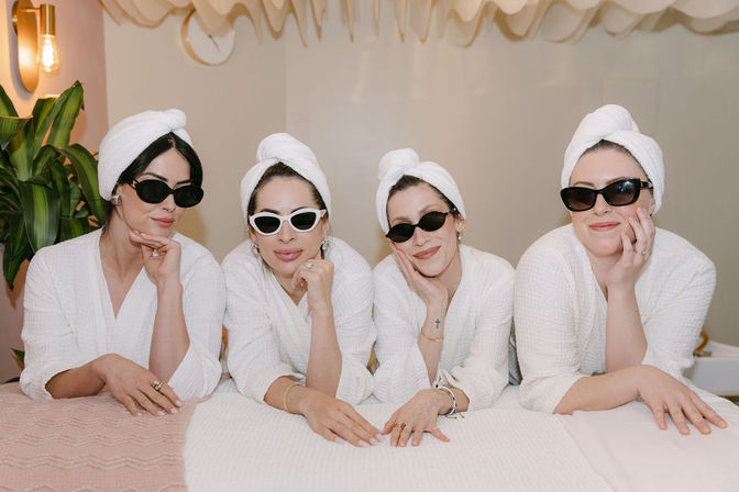 Four women in white robes and towel turbans wearing sunglasses lean on a spa treatment table, smiling in a bright indoor spa room with plants and soft warm lighting.