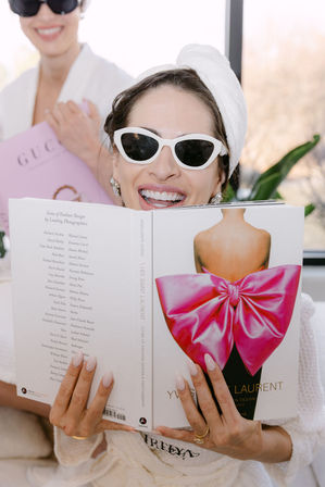 Joyful woman in a towel turban and white cat-eye sunglasses reading a fashion coffee-table book featuring a large pink bow on the cover, sunlit indoor spa vibe with a friend blurred in the background.