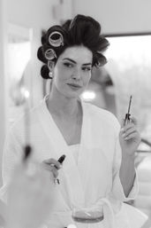 Black-and-white shot of a woman in a white robe with hair rollers, holding a mascara wand at a vanity in a glam getting-ready moment with a drink on the counter.