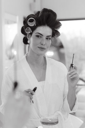 Black-and-white shot of a woman in a white robe with hair rollers, holding a mascara wand at a vanity in a glam getting-ready moment with a drink on the counter.