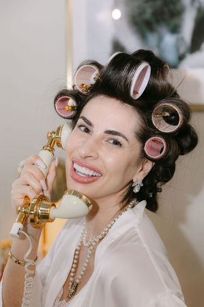 Smiling woman with pink hair rollers and pearl necklace holding a gold-accent vintage phone in a playful getting-ready beauty moment