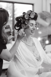 Smiling woman in a robe with vintage hair rollers and a pearl necklace having blush brushed on by a makeup artist during a getting-ready session