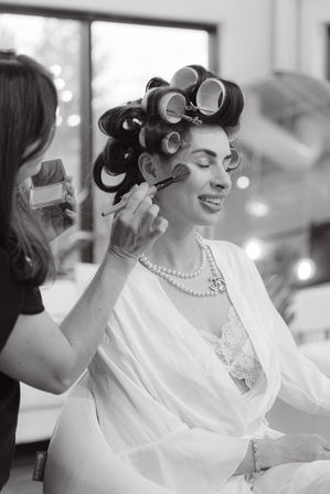 Smiling woman in a robe with vintage hair rollers and a pearl necklace having blush brushed on by a makeup artist during a getting-ready session