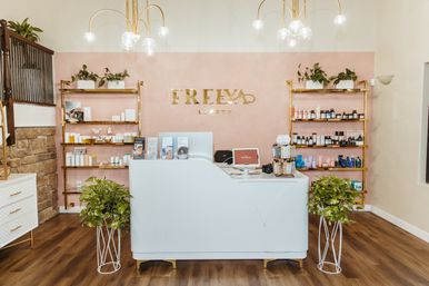 Chic beauty salon reception with white curved desk, pink accent wall, gold shelving stocked with skincare products, potted plants, globe pendant lights and wood flooring.
