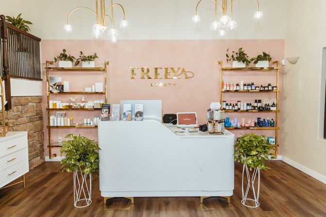 Chic beauty salon reception with white curved desk, pink accent wall, gold shelving stocked with skincare products, potted plants, globe pendant lights and wood flooring.