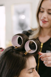 Professional stylist in a beauty salon rolling dark hair into large pink velcro curlers at the front for soft, voluminous curls.