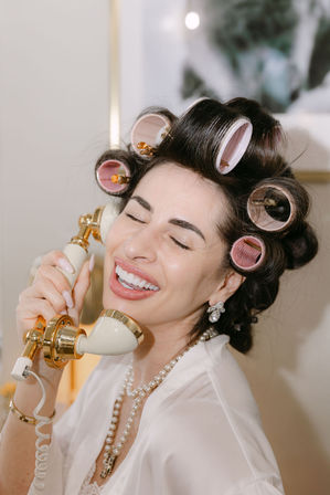 Joyful woman with pink hair rollers and pearl necklace laughing with eyes closed while holding a vintage gold-and-cream telephone handset during a glamorous at-home beauty prep.