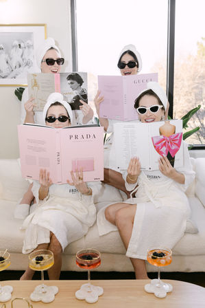 Four women in white spa robes and towel turbans, wearing sunglasses and smiling on a sofa during a playful at-home spa day, reading pink luxury fashion books with cocktails on the coffee table